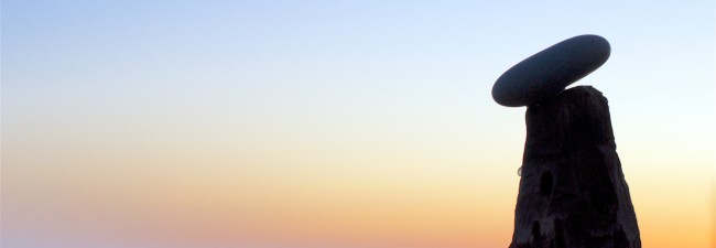 Symbolbild - Ein Kieselstein liegt auf einem angespitzen Baumpfahl. Im Hintergrund sieht man das Meer im Sonnenuntergang
